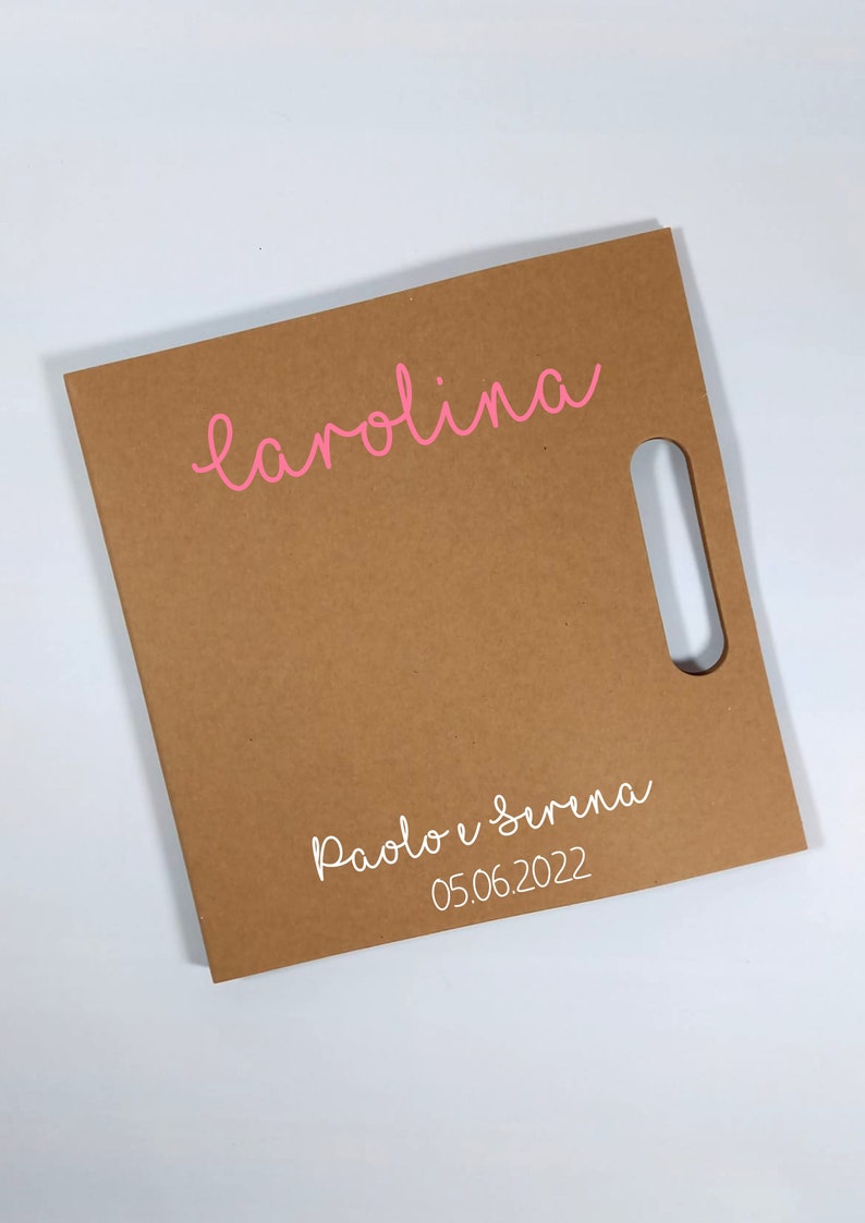 Peut inclure: Un sac cadeau carr&eacute; marron avec une d&eacute;coupe pour la poign&eacute;e. Le nom "carolina" est &eacute;crit en rose en haut, et en dessous, en blanc, les noms "Paolo e Serena" et la date "05.06.2022".