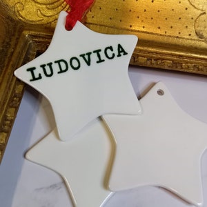 Puede incluir: Dos adornos de cer&aacute;mica blanca en forma de estrella. Un adorno tiene el nombre "Ludovica" escrito en letras verdes.