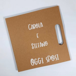 Peut inclure: Un sac cadeau en papier brun avec une d&eacute;coupe rectangulaire pour la poign&eacute;e. Le sac porte le texte blanc "CAROLA E STEFANO OGGI SPOSI". Le sac est sur une surface blanche.