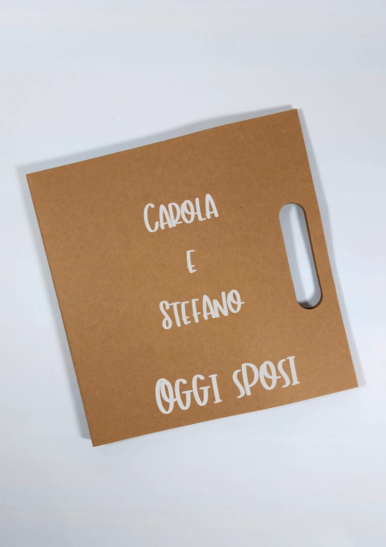 Peut inclure: Un sac cadeau en papier brun avec une d&eacute;coupe rectangulaire pour la poign&eacute;e. Le sac porte le texte blanc "CAROLA E STEFANO OGGI SPOSI". Le sac est sur une surface blanche.