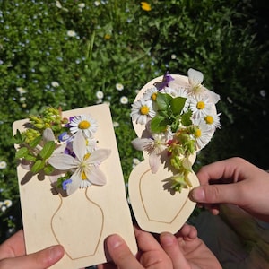 May include: Two wooden vase-shaped cutouts, each decorated with a colourful arrangement of fresh flowers. The flowers include white daisies, purple and white blossoms, and green foliage. The cutouts are held against a backdrop of green grass and wildflowers.