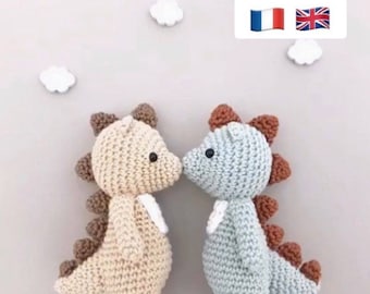 Op de afbeelding: Twee handgemaakte gehaakte dinosaurus speelgoed, een beige en een lichtblauwe, kussen elkaar. Ze hebben bruine stekels en kleine zwarte ogen. De achtergrond is lichtgrijs met drie witte wolkendecoraties.