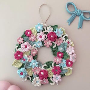 May include: A handmade crochet wreath featuring a variety of colorful flowers in shades of pink, blue, and white, with green leaves. The wreath is circular and has a loop for hanging. A blue bow is in the background.