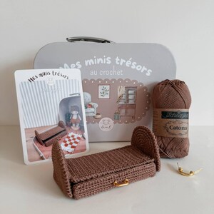May include: A crochet kit with a brown miniature bed, a gray tin with French text, and a ball of brown yarn. The kit includes a small doll and a miniature rug. The tin says "Mes minis trésors au crochet".