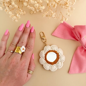May include: A gold-colored ring with a white flower and a small diamond ring are on a hand with pink nails. A crocheted flower-shaped mirror with a gold clasp and a pink bow are also in the image.