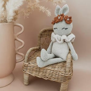 May include: A light grey crocheted bunny doll with a ruffled collar and a flower crown sits on a small wicker chair. The bunny has long ears and closed eyes. The background is a soft, muted pink.