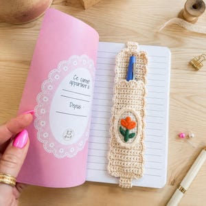 May include: A notebook with a pink cover featuring a white lace-style oval frame with the text Ce carnet appartient à and Depuis. A beige crocheted pen holder with a scalloped edge and an embroidered orange tulip is attached to the lined pages.