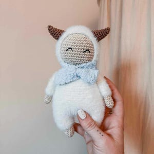 May include: A handmade crochet doll in the shape of a smiling yeti. The doll is white with a fluffy texture, a light blue scarf, and brown horns. The doll has a beige face and is wearing a white hood.