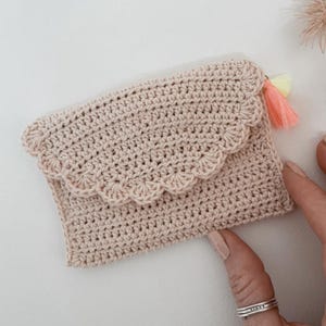 May include: A light pink crocheted wallet with a scalloped edge and a small tassel. The wallet is rectangular with a flap closure. The tassel is a mix of pink and yellow. The crochet stitch is visible.