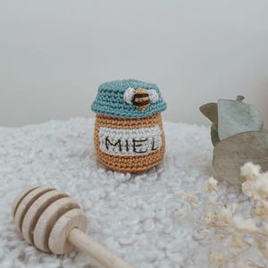 Peut inclure: Pot de miel crocheté avec un couvercle bleu et une petite application d'abeille. Le pot est beige avec le mot "MIEL" brodé sur le devant. Un pot à miel en bois est au premier plan. Le pot est posé sur une surface blanche et moelleuse.