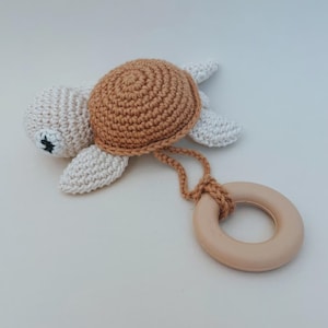 May include: A handmade crochet turtle baby toy in beige, brown, and white. The turtle has a brown shell, white flippers, and a beige wooden ring attached by a brown cord. The toy is designed for babies.