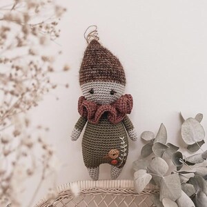 May include: A handmade crochet doll with a brown pointed hat, gray face, and a ruffled burgundy collar. The doll wears a green outfit with a floral design. The doll is displayed on a woven surface.