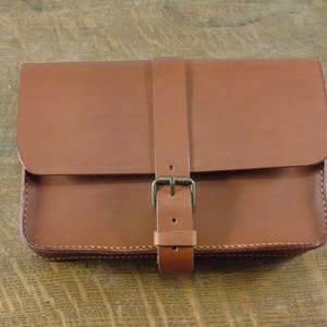 May include: A rectangular brown leather bag with a flap closure and a buckle strap. The bag is made of smooth leather and has visible stitching around the edges. The bag is sitting on a wooden surface.