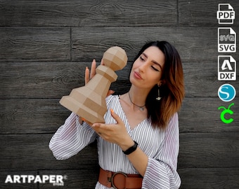 Puede incluir: Una persona sostiene un modelo de peón de ajedrez de papel marrón. El modelo es detallado y tiene una parte superior redondeada. La persona lleva una camisa a rayas y un cinturón marrón. El fondo es una pared de madera con el texto "ARTPAPER" visible en la parte inferior.