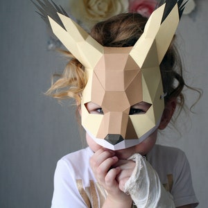 May include: A child wearing a papercraft animal mask with brown and yellow geometric shapes. The mask has large ears and a pointed snout. The child is looking down and has their hands covering their face.