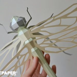 May include: A papercraft dragonfly in shades of green, gray, and beige. The dragonfly has detailed wings and a segmented body. The image includes the text "ARTPAPER STORE" and icons for PDF, SVG, DXF, and other file formats.