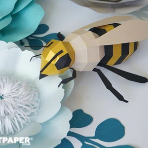 Könnte beinhalten: Eine Papierbiene mit geometrischem Design, mit gelben und schwarzen Streifen, ruht auf einer Papierblume. Die Biene ist von hellblauen und weißen Papierblumen umgeben. Das Bild enthält den Text "ARTPAPER STORE".