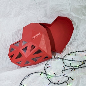 Könnte beinhalten: Eine rote herzförmige Schachtel mit geometrischem Design und einem transparenten Fenster. Die Schachtel ist geöffnet und das Innere ist sichtbar. Die Schachtel steht auf einer weißen Oberfläche mit einer Lichterkette.