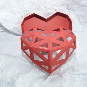 May include: A red heart-shaped box with a geometric design and a clear window. The box is open and the inside is visible. The box is made of paper and has the word "ARTPAPER" printed on the side.