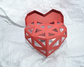 May include: A red heart-shaped box with a geometric design and a clear window. The box is open and the inside is visible. The box is made of paper and has the word "ARTPAPER" printed on the side.