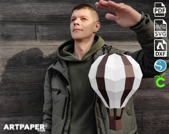 May include: A papercraft model of a hot air balloon with brown and white stripes. The balloon is in focus and the background is blurred. The image includes the text "ARTPAPER" and file format icons for PDF, SVG, DXF, and STL.
