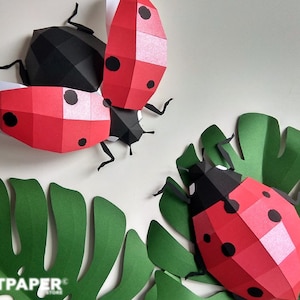 May include: Papercraft ladybug art with red and black segmented bodies and black spots. The ladybugs are positioned near green paper leaves. The image includes the text "ARTPAPER STORE".
