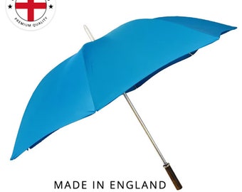 Puede incluir: Un paraguas azul brillante con un mástil plateado y un mango de madera marrón oscuro. El paraguas está abierto, mostrando su capota. El texto "MADE IN ENGLAND" está impreso debajo, con un emblema circular encima.