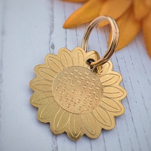 May include: A gold-coloured sunflower-shaped dog tag with a ring for attaching to a collar. The tag has a detailed sunflower design with a textured centre.