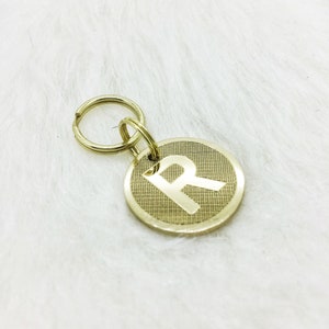 May include: Gold-colored metal dog tag with a round shape and a single letter "R" engraved on the front. The tag has a split ring attached to it.