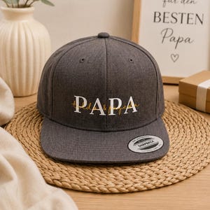 Könnte beinhalten: Eine anthrazitfarbene Snapback-Cap mit dem Wort PAPA, das in weißen Großbuchstaben mit einem dünnen goldenen Schreibschrift-Overlay gestickt ist. Der flache Schirm hat einen runden schwarz-weißen Aufkleber mit der Aufschrift The Authentic Snapback.