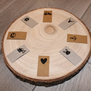 Könnte beinhalten: Eine runde Holzscheibe mit acht kleinen rechteckigen Etiketten mit verschiedenen schwarzen Symbolen darauf. Die Symbole beinhalten einen Traktor, ein Reh, einen Anker, eine Blume, ein Herz, ein Kaninchen, eine Schaufel und einen Kreis mit einem Strich durch.