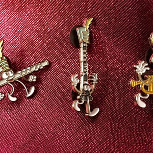 May include: Three enamel pins featuring cartoon characters playing musical instruments. The characters are wearing a marching band hat and have a white face with black eyes. The instruments are a flute, a clarinet, and a trumpet. The pins are on a red background.