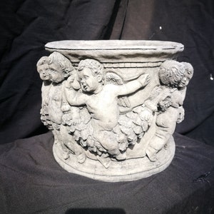 Könnte beinhalten: Ein grauer Betontopf mit einer runden Oberseite und einem Design von Cherubim mit Flügeln an den Seiten.