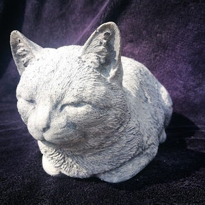 May include: A grey concrete cat sculpture with a realistic design. The cat is lying down with its head turned to the side and its eyes closed. The sculpture is made of a rough, textured material.