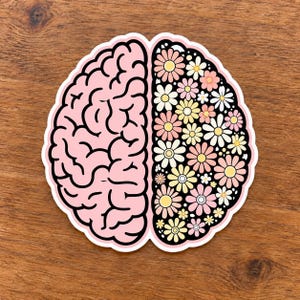 May include: A die-cut sticker shaped like a human brain. The left hemisphere is solid light pink with black outlines representing the sulci. The right hemisphere is black and filled with a pattern of white, yellow, and pink daisy flowers.