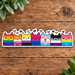 May include: A horizontal sticker featuring seven cartoon cats arranged in a row, each colored with different pride flag patterns including rainbow, lesbian, non-binary, bisexual, transgender, asexual, and pansexual, topped with small hearts.