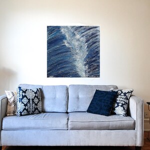May include: A gray sofa with decorative pillows in shades of blue and white. Above the sofa hangs a square abstract painting in shades of blue and white. A small black side table with a gold base is to the right.