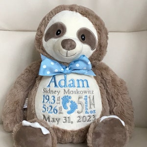 Peut inclure: Une peluche paresseux marron avec un nœud papillon bleu clair à pois blancs. Le ventre est brodé avec le nom Adam, Sidney Moskowitz, les détails de naissance 19.3 inches, 5 pounds 14 ounces, 5:26 pm, le 31 mai 2023 et des empreintes de pieds bleues.