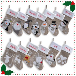 May include: A selection of Christmas stockings with names embroidered in red on white cuffs. Each stocking features a festive design, including penguins, snowmen, reindeer, and a paw print. The stockings are beige with snowflake accents.