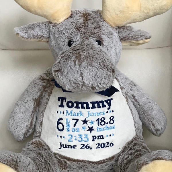 May include: A plush gray moose stuffed animal with tan antlers and a dark blue ribbon around its neck. The white belly features embroidered text: Tommy, Mark Jones, 6 lbs 7 oz, 18.8 inches, 2:33 pm, and June 26, 2026, with blue stars.
