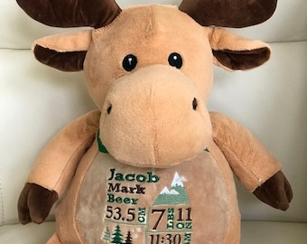 May include: A plush tan moose toy with dark brown antlers and hooves. The chest features embroidered text: Jacob Mark Beer, 53.5 cm, 7 lbs 11 oz, 11:30 AM, and October 9, 2024, alongside green mountain, tree, and arrow graphic icons.