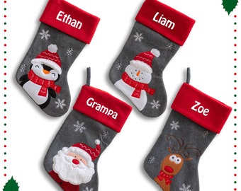 May include: Four gray Christmas stockings with red cuffs, each personalized with a name. The stockings feature embroidered designs of a penguin, snowman, Santa Claus, and a reindeer. Snowflake accents are also present.