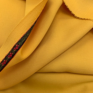 May include: Close-up of a yellow fabric with a red and black trim. The fabric is textured and has a subtle sheen. The trim has a geometric pattern. The fabric is labelled "ITALY".