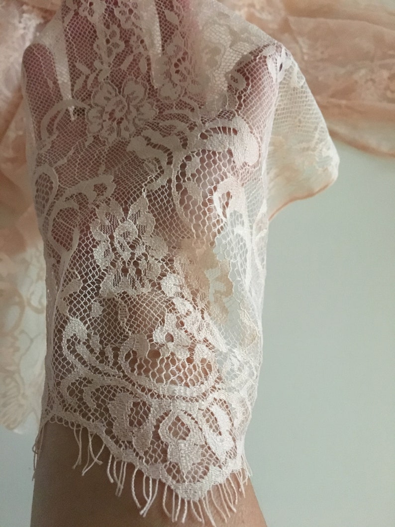 May include: A close-up of a delicate, pale pink lace fabric with a scalloped edge. The lace has a floral pattern and a soft, airy texture.