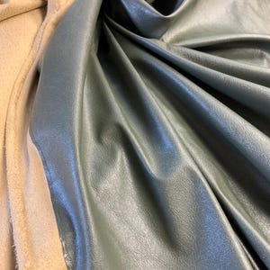 May include: Close-up of a dark green faux leather fabric with a smooth, slightly textured surface. The fabric is folded and draped, showcasing its soft, pliable texture.