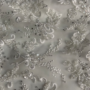 Puede incluir: Tela de encaje blanca con delicado bordado floral y cuentas brillantes.