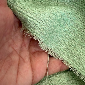 Può includere: Primo piano di un tessuto strutturato verde chiaro con un motivo intrecciato. Il tessuto ha una frangia lungo il bordo e un filo sciolto. Il materiale sembra essere un tweed o un tessuto simile.