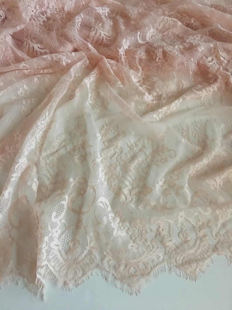 May include: A piece of delicate, off-white lace fabric with a subtle ombre effect, transitioning from a light pink to a white hue. The lace features a floral pattern and a scalloped edge.