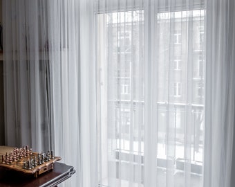 May include: White sheer curtains hang in front of a window with a view of a snowy cityscape. The curtains are gathered at the top and hang in soft folds. A wooden table with a chess set sits in the foreground.