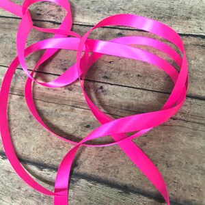 May include: A close-up of a pink satin ribbon on a wooden surface. The ribbon is folded and twisted, creating a decorative pattern.